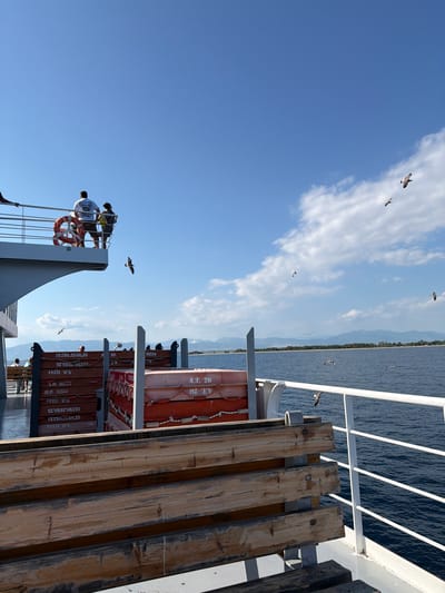 The image depicts a sunny day with a clear blue sky and scattered white clouds, viewed from the deck of a ferry in Keramoti, Greece. The ferry is in motion, with the dark blue sea visible on the right side of the frame. On the upper deck, two individuals stand near a railing, one facing away from the camera. A life preserver ring is attached to the railing. In the middle ground, stacked orange life rafts are visible. Several seagulls are in flight across the sky and above the water. The background shows a distant coastline with a line of trees and mountains under the horizon. In the foreground, wooden benches are positioned on the ferry deck.