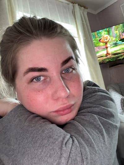 The media file is a close-up, tilted, vertical selfie of a young person lying down and looking at the camera. The subject has fair skin with freckles, blue eyes, and dark eyebrows. They are wearing a grey hooded sweatshirt. The background shows a television screen displaying a cartoon with a brown creature and a green, leafy environment, suggesting the person is in a living room or bedroom. Natural light is coming in from the left side, illuminating the person's face and casting shadows. The overall mood appears relaxed and casual. There is no visible text in the image. The location context is Уральское, Russia.