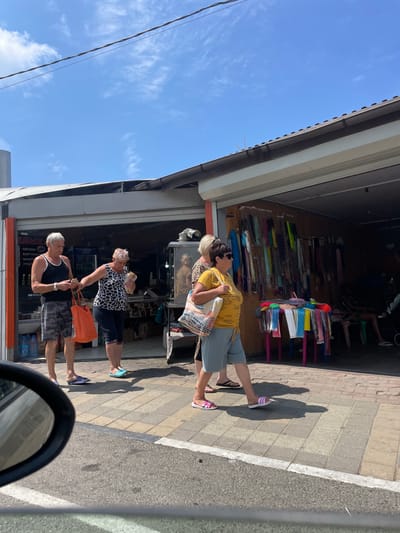 In Dzhubga, Russia, on a bright, sunny day, a scene unfolds along a roadside marketplace. Four individuals, two men and two women, are captured in a candid moment. The foremost man, clad in a black tank top and camouflage shorts, carries an orange tote bag in his left hand and appears to be holding something small in his right. To his right, a woman in a black and white patterned sleeveless top and dark shorts is extending her right hand, possibly interacting with a small white bag she holds. Behind her, a woman with short blonde hair, wearing a yellow t-shirt and light blue shorts, walks with a patterned tote bag slung over her shoulder. Another woman, with short dark hair and sunglasses, stands to the left of the yellow-shirted woman, also with a tote bag. The backdrop is a storefront featuring an open-air market with various items displayed, including colorful textiles and what appear to be food items. The ground is paved with interlocking square tiles. The overall atmosphere suggests a casual daytime outing in a commercial area.