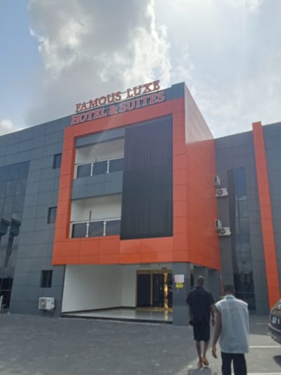 The image shows a hotel building located at 5.472, 5.817 in the city of Ujevwu, Nigeria. The building is three stories tall and has a modern design. The facade of the building is orange and gray. The hotel sign reads "Famous Luxe Hotel & Suites" in red lettering. The building's entrance is located on the ground level, and there is a large glass door with a golden trim. There are two men walking towards the entrance. The sky is bright blue with white clouds.