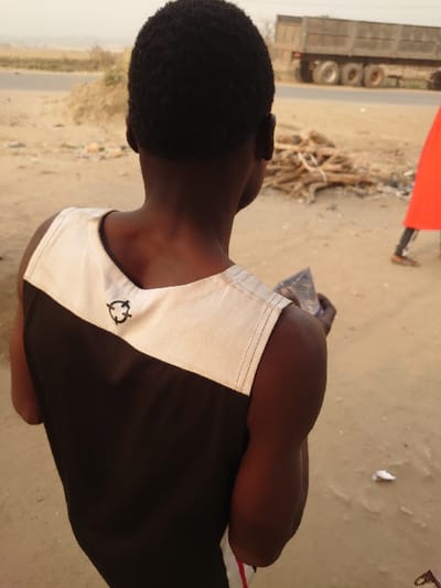The image presents an outdoor scene in Ajaokuta, Nigeria, characterized by dry, dusty conditions. In the foreground, a person with dark skin and short, dark hair is depicted from the back, slightly over their right shoulder. This individual wears a sleeveless garment that is predominantly black, with a cream or light tan upper panel featuring a black, stylized circular emblem near the left shoulder blade. Their right arm is bent, holding an indistinct dark, crumpled object.
The person stands on uneven, light brown, dusty terrain. In the midground, a paved road extends horizontally across the frame. Adjacent to the road, a pile of stacked wooden branches or logs is visible. To the far right, a second individual's lower body is partially visible, clad in a bright red garment and brown footwear.
In the background, a large, multi-wheeled grey heavy-duty truck, possibly a dump truck, is situated on the paved road. The distant horizon appears hazy, consistent with atmospheric dust or a far-off landscape. The overall lighting suggests daylight.