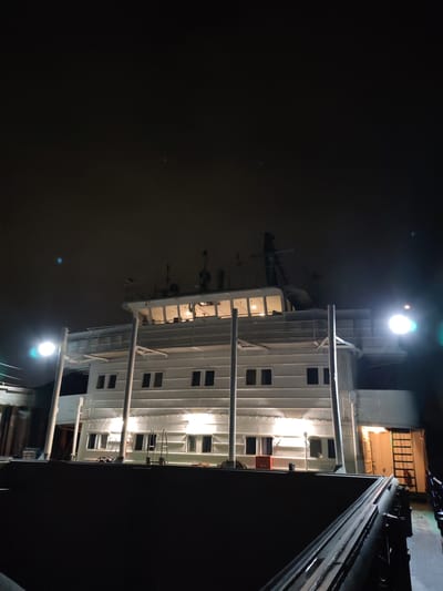 The image depicts a large, illuminated vessel at night. The vessel has multiple levels, visible windows, and supporting columns. It's illuminated by bright lights. The sky is dark, and the scene appears to be on the water, given the context of Tver, Russia. It's plausible that the vessel is on the Volga River in Tver.