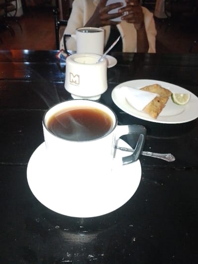 A steaming cup of coffee sits on a white saucer, positioned on a dark, glossy table. A white sugar bowl with a spoon and a filled mug are in the background. A white plate holds two triangular pastries, one wrapped in a napkin, with a lime wedge. A silver spoon rests beside the coffee cup. An individual in a beige jacket is visible in the blurred background, focused on a phone.