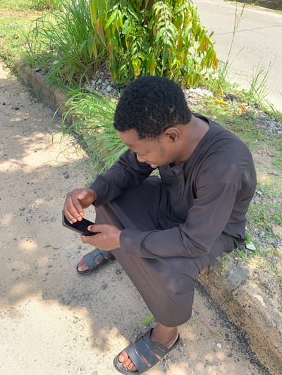 A person, appearing to be a young Black male, is crouched down outdoors, looking at a smartphone. He is wearing a dark, possibly black, tunic and dark sandals. The ground beneath him is a mix of dirt and small gravel, with patches of green grass and some leafy plants to his left. A road is visible in the background. The lighting suggests it is daytime, with shadows indicating sunlight. The scene appears casual and perhaps contemplative, given the subject's focus on the phone and his relaxed posture. No visible text is present on the clothing or objects. The image seems to capture a moment of everyday life in a rural or semi-rural setting, possibly in Galadimawa Village, Nigeria.