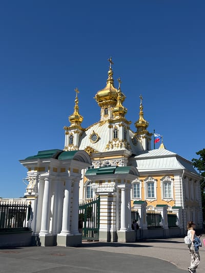 The image depicts the Peterhof Palace, specifically the Church of the Icon of the Mother of God Joy of All Who Sorrow, with its striking golden domes against a clear blue sky. The building exhibits Baroque architecture, characterized by ornate gold embellishments, white columns, and yellow facades with decorative window frames. A Russian flag is visible atop one of the domes.

In the foreground, a person in casual clothing, wearing a white top and patterned pants, is looking at their phone, leaning against a low wall. The lighting suggests it is daytime, with the sun casting distinct shadows from the building's architectural features and the nearby trees, indicating a sunny and pleasant weather condition. The overall scene is one of historical grandeur and a peaceful, perhaps tourist-filled, atmosphere.