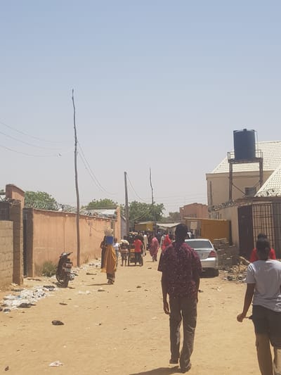 The image captures a street scene in Dabawa, Nigeria. Several individuals are present, including a man in a maroon patterned shirt and gray pants walking on the right side of the frame, and another individual in a light-colored shirt standing near a vehicle. In the background, a group of people appear to be gathered, some possibly carrying items. The environment consists of buildings with barbed wire fencing and overhead utility lines. There is a water tank atop one of the buildings. A motorcycle is parked near a wall. The ground is sandy, and there is visible trash scattered along the street.