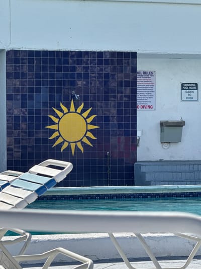A daylight scene depicts a swimming pool area located in Hallandale Beach, United States. The background features a wall segmented into two sections. The left section is tiled with dark blue square tiles, centrally displaying a large yellow sun mosaic. Above the sun, a wall-mounted shower head is visible, with a small pipe or drain below it. The right section of the wall is painted white, featuring two regulatory signs. The larger sign, titled "POOL RULES," lists "HOURS 7AM TO 10PM" and includes a "NO DIVING" warning in red text. Adjacent to it, a smaller sign indicates "SWIMMING POOL HOURS DAWN TO DUSK." Below these signs, a beige, wall-mounted water fountain is present, situated above a grey concrete structure with steps. The foreground reveals a swimming pool with clear, blue-green water and a blue tiled coping. Partially visible in the mid-ground is a white and light blue striped lounge chair, and a white metal railing extends across the immediate foreground. No individuals are present within the frame.