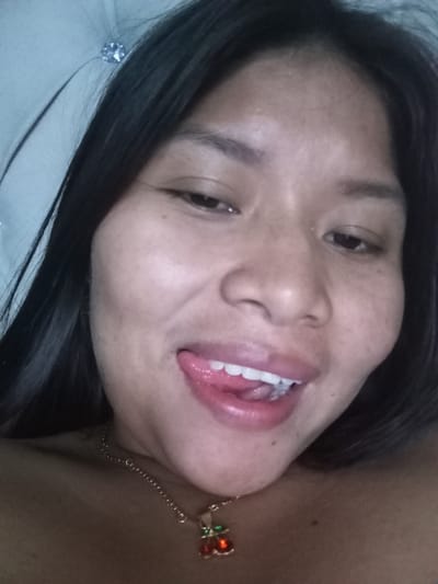 A woman takes a close-up selfie, her dark hair cascading around her face. She has a cherry necklace. Her eyes are dark, and she playfully sticks her tongue out. Her skin is smooth. She is laying down. In the upper left is a tufted headboard with a crystal button.