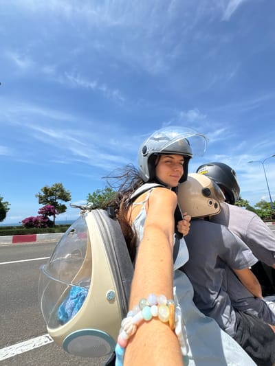 In Mui Ne, Vietnam, three people ride a motorcycle. The person in the foreground, a woman with long, dark hair, wears a silver helmet and a light blue top. Her arm, adorned with a beaded bracelet, extends forward, holding the camera. Behind her, another woman, wearing a tan helmet, and a third person, in a black helmet, are also on the motorcycle. A cream-colored, clear plastic pet carrier is attached to the back of the motorcycle. The background shows a road with a red and white border, lush green trees, and vibrant purple flowers under a bright blue sky with scattered clouds.