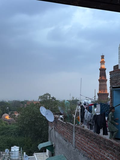 The image depicts a view of the Qutub Minar in Delhi, India, under a cloudy sky. In the foreground, a brick wall runs along the edge of a rooftop. Clothes are hung on a clothesline on the rooftop, with several satellite dishes visible. A young woman in a green outfit is walking on the rooftop, facing away from the camera and towards a blue door. In the background, lush green trees obscure some of the lower structures. The Qutub Minar, a UNESCO World Heritage Site, stands tall in the distance, illuminated and prominently visible against the hazy sky. Another domed structure is partially visible amongst the trees to the left. The overall scene suggests a residential area in close proximity to historical landmarks.