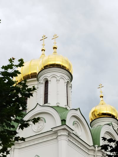 This media file features a close-up, low-angle shot of several golden domes and green roofs of an Orthodox church, set against a cloudy, overcast sky. The architectural style is characteristic of Russian churches, with onion-shaped domes topped by crosses. The main subjects are the church's domes and upper structures, prominently displaying their gilded surfaces and white stonework. Green foliage from a tree in the foreground partially obscures the lower left portion of the image, adding a natural element to the scene.
The weather appears to be cloudy, indicated by the uniform grayness of the sky, which suggests it could be morning, afternoon, or evening on a day with limited sunlight. The image captures a static scene, focusing on the building's impressive architecture rather than an ongoing activity or event.
Notable details include the intricate design of the crosses on the domes, the arched windows visible on the church's facade, and the decorative relief work on the white sections of the building. The golden domes gleam despite the muted lighting, highlighting their importance as a focal point. The presence of a tree suggests the church is situated in an area with some greenery.
No people are visible in the frame, and there is no discernible text. The overall impression is one of grandeur and religious significance, conveyed through the imposing structure and its ornate features. The location context of Pushkin, Russia, is a key piece of information that helps identify this type of architecture.