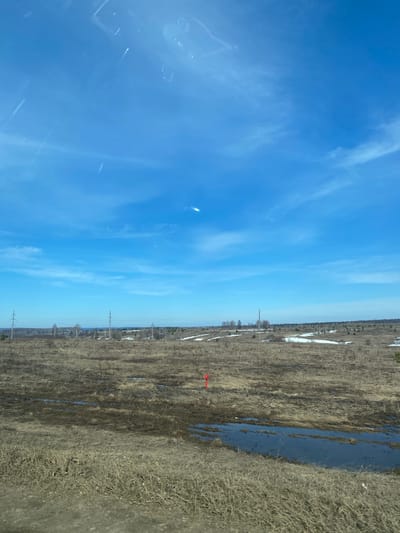 The image shows a landscape in Kueda, Russia. The sky is a clear blue with scattered cirrus clouds. Below the sky is a rural landscape with a field partially covered in dried vegetation and patches of melting snow. Power lines stretch across the horizon. A narrow stream or standing water runs through the field. A red arrow is superimposed on the image in the center of the field.