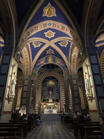 This image depicts the interior of the Basilica di Santa Margherita, located in Cortona, Italy. The view is from the nave, looking towards the main altar at the far end of the church.

The environment is a grand, high-ceilinged basilica characterized by ornate Neo-Gothic and Romanesque revival architecture. The vaulted ceiling is painted a deep blue, adorned with numerous gold stars and intricate geometric and floral patterns in red, gold, and white along the intersecting arches. Several circular and star-shaped medallions containing depictions of religious figures and angels are embedded within the ceiling and archways.

Large, structural pillars line both sides of the central aisle, featuring alternating vertical bands of red-brown and cream-colored stone or paint. White marble statues, depicting robed figures, are mounted on pedestals along these columns. The central nave features a dark and light checkered floor pattern, likely tile or stone, leading towards the altar.

The sanctuary, located at the far end, is elevated by several steps. It contains a white altar table and a decorated reredos behind it. A large, ornate chandelier hangs directly above the altar, and a prominent gilded crucifix or monstrance is centrally positioned on a stand. The rear wall of the sanctuary features arched elements, including an organ.

Approximately 15-20 individuals are visible, primarily seated in wooden pews arranged in rows on both sides of the central aisle. Most are facing towards the altar. Their actions are largely static; they appear to be attending a service or engaged in quiet contemplation. One individual in the immediate foreground on the right is partially visible, facing away from the camera. No direct interactions between individuals are clearly discernible.