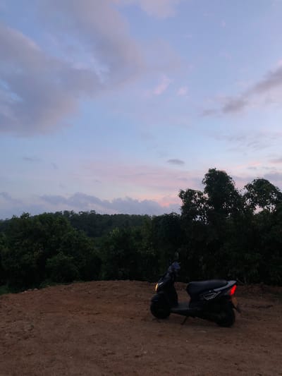 A black scooter is parked on a dirt mound overlooking a lush green forest in Kathaluwa, Sri Lanka. The sky above is a pastel gradient of blue, pink, and purple, with soft clouds drifting by. The scene appears to be set during dusk or dawn. The scooter is positioned to the right of the frame, facing left. Its headlights are illuminated, casting a warm glow on the dirt path. The forest stretches across the background, a dense expanse of trees. The ground beneath the scooter is dry and sandy, with small rocks scattered throughout. The overall atmosphere is serene and natural.