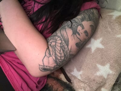 A close-up shot depicts a person's tattooed leg resting on a beige pillow with white stars, partially covered by a pink blanket. The tattoo features a woman's face and a snake-like creature with what appears to be scales. A strand of dark hair lies between the leg and the blanket. The location cannot be determined from the image.