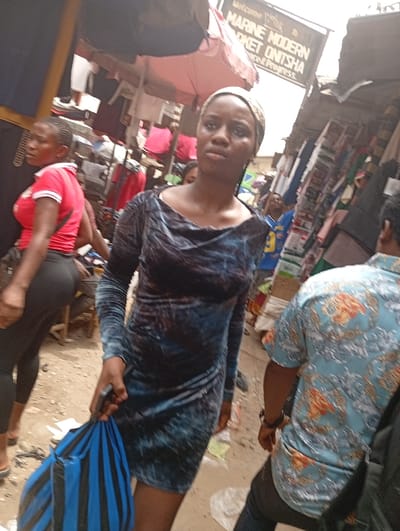 A young woman in a blue, velvet-like dress with long sleeves and a draped neckline stands in a bustling market. She wears a headscarf and holds a blue and black striped bag. Behind her, other people walk through the crowded area, including a woman in red and black athletic wear and a man in a patterned shirt. Market stalls and signage are visible, indicating a busy outdoor commercial environment. The sign reads, "Welcome to Marine Modern Market Onitsha."