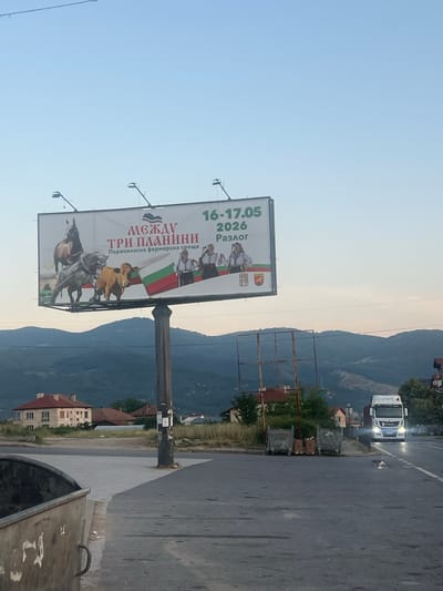 The image shows a billboard advertising an event called "МЕЖДУ ТРИ ПЛАНИНИ" (Between Three Mountains), which is identified as "Първокласна фермерска среща" (First-class farmer's meeting). The dates for the event are listed as "16-17.05 2026" in Razlog. The billboard features images of horses, a cow, and people in traditional Bulgarian attire, along with the Bulgarian flag.
In the foreground, a large metal dumpster is visible on the left side of the frame. The billboard is supported by a tall, weathered pole. Behind the billboard, rolling hills and mountains are visible, with some houses and buildings scattered across the landscape. A white truck is driving on a road in the middle ground of the image, with its headlights illuminated. The sky is clear and pale blue, suggesting it is either early morning or late afternoon. The overall scene depicts a rural or semi-rural setting.
The location is identified as Simitli, Bulgaria, based on the context of the "Between Three Mountains" event, which is known to take place in or around that region of Bulgaria.