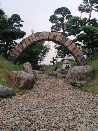 The image depicts a scenic pathway in a park located in Hong Gai Ward, Vietnam. A stone archway frames the entrance to the path, which is covered in small, rounded stones. Large boulders flank the path on either side, leading into a landscaped area with grass and trees. A traditional Japanese-style stone lantern is situated near the archway, providing light. In the background, bamboo stalks and additional signage can be observed. The sky is overcast.