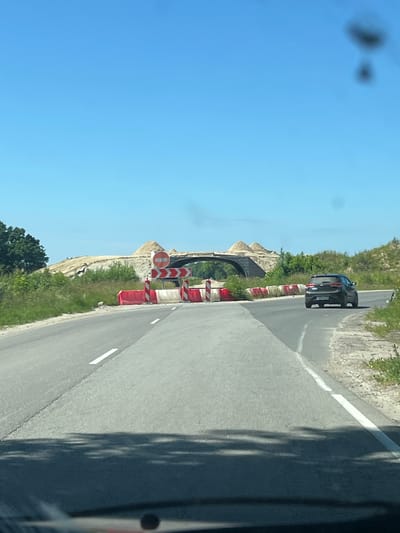 In Putilovo, Russia, a road curves to the right, leading towards an under-construction overpass. The overpass is partially built with mounds of sand and dirt, and a dark, arched structure is visible underneath. Red and white barrier blocks line the road, indicating a construction zone. A dark gray sedan is driving away from the camera on the right side of the road. The sky is clear and blue, and green trees and vegetation are visible on the sides of the road and around the construction site. The road is asphalt and has white lane markings. Shadows of trees are cast on the road in the foreground, suggesting the photo was taken from a vehicle.