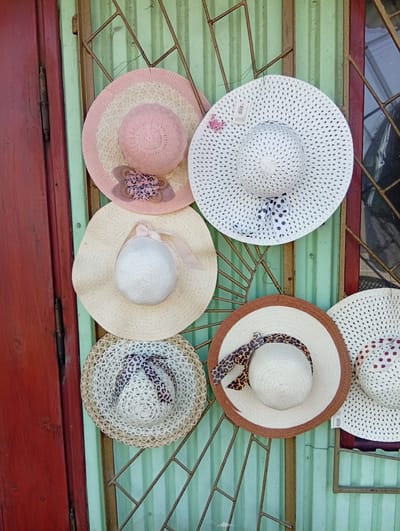 Six straw hats are displayed on a green corrugated metal wall, hanging from a metal grate. The hats are of various designs and colors, including a pink and white striped hat with a floral embellishment, a white hat with a black polka dot ribbon, a light beige hat with a sheer pink ribbon, a white hat with a leopard print ribbon, and a white hat with a brown band and a leopard print ribbon. The wall is next to a red wooden door and a window with a metal grate. The scene appears to be an outdoor market stall in Długołęka, Poland.
