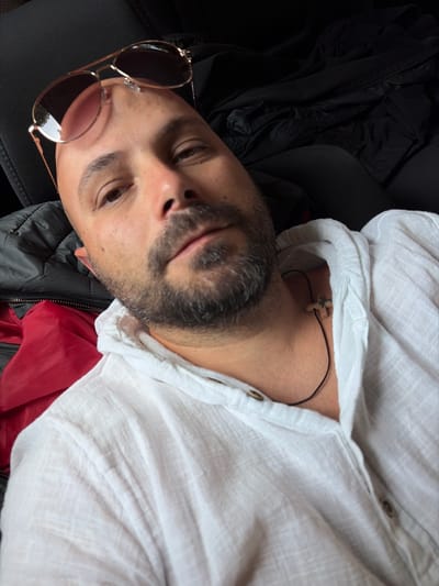 A man is lying down inside a vehicle. He is wearing a white collared shirt with the top buttons undone and a thin black cord necklace with a wooden cross pendant. He also has on aviator-style sunglasses with rose-tinted lenses perched on his bald head. The man has short, graying stubble on his face. In the background, a red garment and a black jacket are visible. The scene is likely in Cherven Bryag, Bulgaria, based on the provided context.