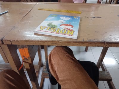 A wooden desk with a brown surface is in focus. There is a book titled "NOISIA" lying on the desk. It is open, but the pages are not visible. A yellow ruler lies on the desk near the book, partially visible. On the left side of the desk, part of a pair of brown pants is seen hanging over the edge. The desk is resting on two wooden legs, which are visible on the left and right sides of the image. The desk is in a classroom, as seen in the image background. The location is Batu, Indonesia, coordinates -7.879, 112.527. The scene is otherwise empty.