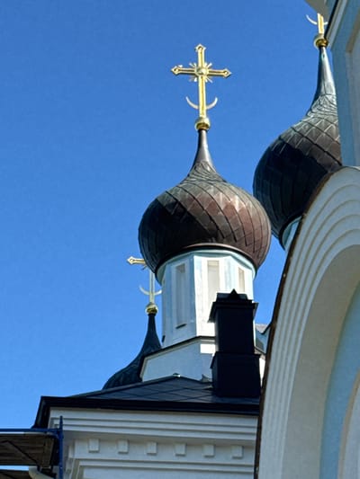 The image shows the domes and crosses of a Russian Orthodox church, seen against a clear blue sky. The architecture features multiple onion domes, one of which is a prominent dark, metallic color, while another is lighter and features arched architectural elements. Golden crosses, characteristic of Orthodox churches, are mounted atop the domes. One cross is particularly detailed with a circular element and a crescent moon shape at its base. The church building itself appears to be white with light blue accents. A black chimney or vent is visible on one of the structures. The scene is a detail-oriented shot, focusing on the religious architecture. The location is identified as St. Ozerki, Russia. There are no people or animals depicted in the image.