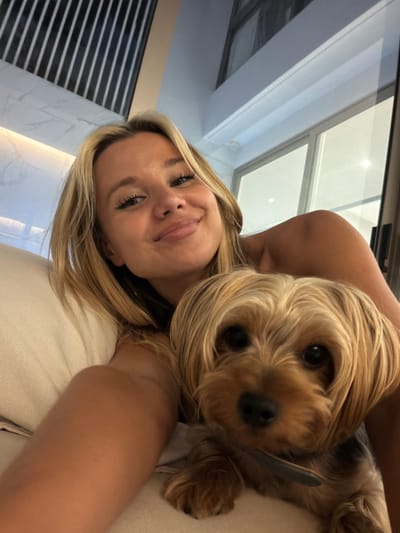 In an indoor setting, a blonde woman with blue eyes and a smile is taking a selfie with her Yorkshire Terrier. The woman is lying down, with her head propped on a light-colored cushion. The dog is in the foreground, looking directly at the camera. The background appears to be a modern interior with a large window and vertical wooden blinds on a wall. The overall scene is casual and intimate, suggesting a relaxed moment at home. The specific location in Thep Krasatti, Thailand, is not discernible from the visual information provided.