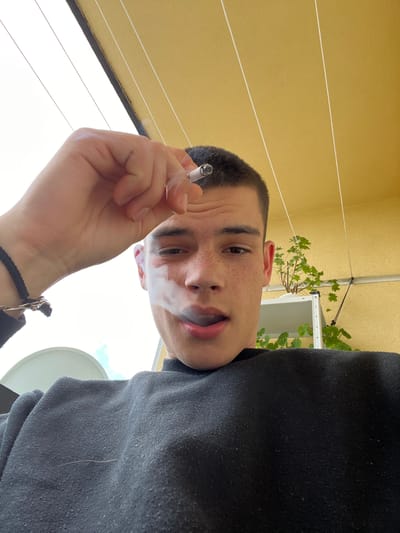 A close-up, low-angle shot features a young adult male with fair skin, short dark hair, and visible freckles. He is holding a lit cigarette in his right hand, exhaling smoke. He is wearing a dark grey or black sweater. A black braided bracelet is on his wrist.
The background includes a yellow wall with horizontal lines and some greenery. A white satellite dish is visible in the lower right corner of the frame. The location is not in Bulgaria and cannot be proven to be Sopot.