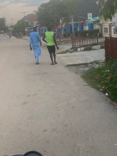Two people walk along a paved street in Iddo Tudunwada, Nigeria. One person wears a light blue outfit, while the other wears a neon green shirt and black shorts. Buildings and trees are visible along the side of the street. Power lines stretch above the street.