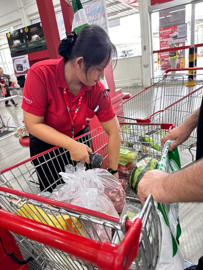 In Wichit, Thailand, a female store employee wearing a red polo shirt with the "CP TRA" logo is scanning items in a shopping cart. The cart contains various groceries, including a watermelon, celery, tomatoes, and several plastic bags of food, possibly meat. In the background, a person in a high-visibility vest is visible near a checkout counter, and other shoppers can be seen in the distance. The environment appears to be a brightly lit supermarket or large retail store, indicated by the rows of shopping carts, overhead lighting, and signage. A customer's hand is reaching into the cart to place a bag of groceries.