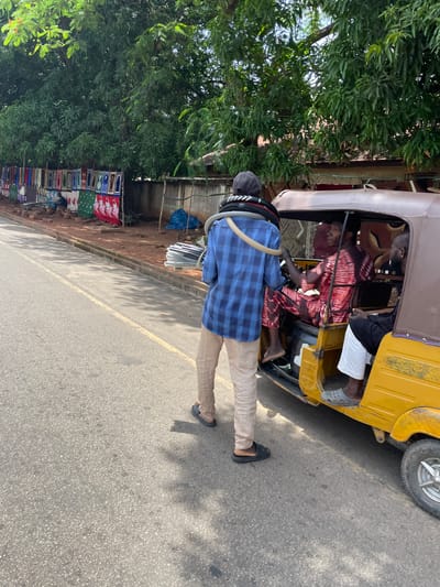A medium shot captures a street scene in Unguwar Sarki, Nigeria, with a bright yellow auto-rickshaw parked on the right side of the frame. A man wearing a blue checkered shirt and beige striped pants stands by the open door of the auto-rickshaw, carrying a coil of black and white tubing over his left shoulder. Inside the auto-rickshaw, a driver in a red and white patterned dashiki is seated at the controls, looking forward. Another person, wearing white pants, sits in the passenger seat. To the left of the auto-rickshaw, a row of stalls displays various textiles and fabrics against a low wall. The ground is a paved road with a yellow lane marker, and a dirt shoulder runs alongside it, lined with a curb. Lush green trees with dense foliage arch over the scene, creating dappled shade on the road. The overall impression is of a busy roadside market on a sunny day.