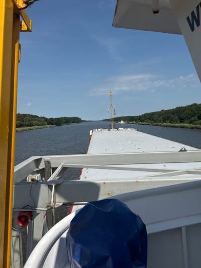 This image captures a sunny day from the deck of a large vessel navigating a calm body of water, likely a canal, bordered by lush green trees. The perspective is angled, looking down the length of the ship. The foreground features a portion of the vessel's deck, with yellow railing and part of a white structure with black lettering visible on its side. Further along the deck, a wide, flat, grey surface with numerous evenly spaced dots stretches ahead. A tall mast with flags is visible in the mid-ground. In the background, another vessel is partially seen. The water is a deep blue, reflecting the clear sky. A large blue tarp covers an object on the right side of the frame, and a red horn or speaker is mounted on a grey metal structure. The presence of the canal and the German location context suggest this might be the Kiel Canal. The bright sunlight indicates it's daytime, likely mid-morning to afternoon, with clear skies and fair weather.