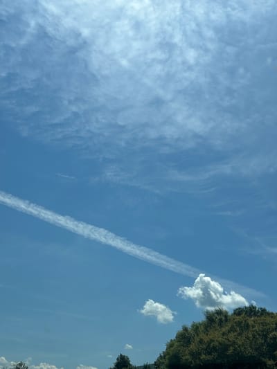 The image displays a clear blue sky with wispy clouds and a prominent contrail stretching diagonally across the upper left quadrant. In the lower right corner, the top of a lush green tree line is visible, suggesting an outdoor environment. Two small cumulus clouds are present above the trees. The image appears to be taken from a ground-level perspective, looking upwards. Based on the provided information, this scene is located in the city of Lozen, Bulgaria.