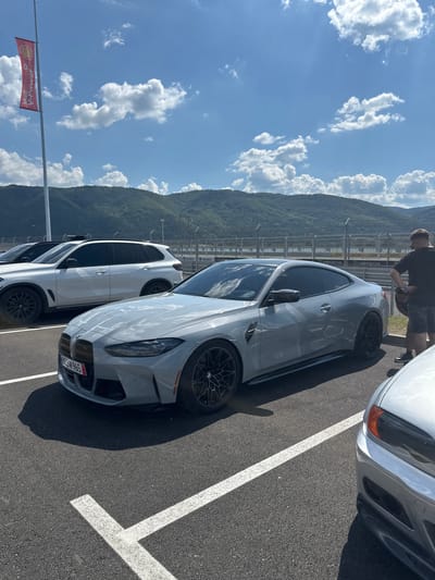 A light gray BMW M4 coupe is parked in a paved lot with white parking lines, facing right. The car has a red license plate with white lettering that reads "BS3 M965". In the background, a white BMW SUV is parked behind the coupe. Further back, a man in a black t-shirt and shorts is standing near the BMW M4, looking down. A red and yellow Shell V-Power flag on a tall pole is visible to the left, and a chain-link fence with advertisements lines the background. Rolling green hills and mountains under a blue sky with scattered white clouds form the backdrop. The scene appears to be a racetrack or event venue, possibly in or near Dospey, Bulgaria, given the mention of the location.