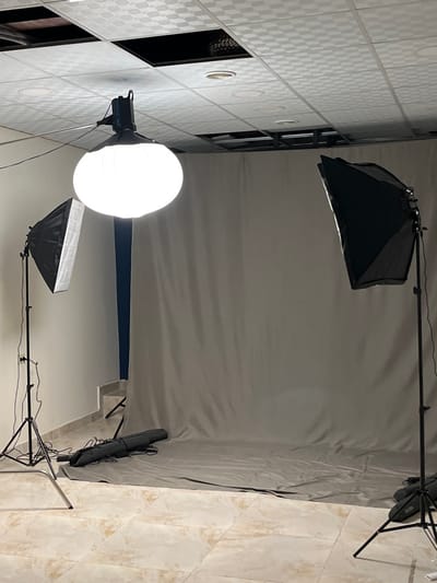 The scene depicts an unoccupied indoor photography studio setup within Al-Mazza Municipality, Syria. The room features a white drop ceiling with several missing panels, exposing dark structural elements and utilities above. Recessed circular lights are present in the ceiling, some illuminated. A large, illuminated spherical light diffuser is suspended centrally from the ceiling. Two additional photography light stands, each fitted with a rectangular black softbox diffuser (white interior), flank a seamless gray fabric backdrop that covers the rear wall and extends onto the floor. The left light stand is positioned near a corner where a dark blue vertical stripe is visible on the wall. The floor is covered with light beige square tiles laid in a diagonal pattern. Dark, rolled equipment bags or cases are visible on the floor near the bases of both light stands. No individuals are present in the image.