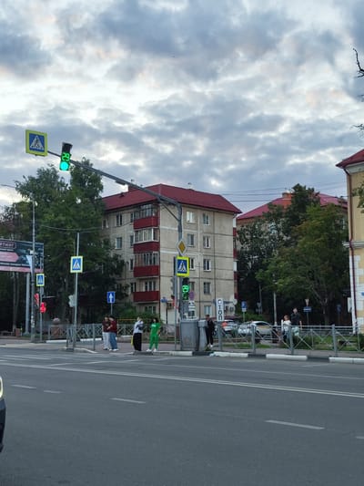 A street scene in Ramenskoye, Russia, with a crosswalk and traffic lights. Several people are visible at the crosswalk, waiting to cross or in the process of crossing. Two traffic lights display green, indicating it is safe to proceed. A pedestrian crossing sign is present. Buildings line the street, some with red roofs. The sky is cloudy, suggesting overcast weather. The time of day appears to be late afternoon or early evening. There is some visible text on signs, but it is mostly illegible or too small to read clearly. One sign, partially visible, reads "СТОП" (STOP) in Cyrillic.