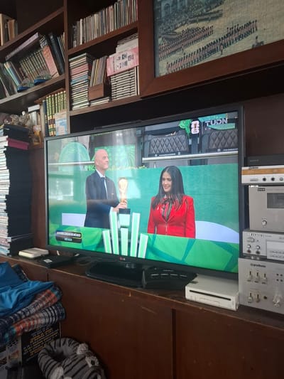 The image depicts an indoor scene, likely a living room or entertainment area, focused on a television screen displaying a news broadcast. The television shows two individuals on a stage with green and white geometric graphics. On the left, a man in a dark suit is holding a trophy. On the right, a woman in a red jacket and a green and red patterned scarf is smiling. The broadcast appears to be from TUDN and mentions "Estadio Ciudad de México" and "México."

To the left of the television, a bookshelf is filled with various books and what appear to be DVDs or CDs. Below the television, a white gaming console, possibly a Nintendo Wii, is visible along with several electronic audio devices stacked vertically. In the foreground, a blue blanket and a patterned throw are draped over furniture, and a grey plush slipper is on the floor.

The ambient environment suggests a home setting with a cluttered but organized collection of media and electronics. The location is identified as Naucalpan de Juárez, Mexico, based on information on the television screen.