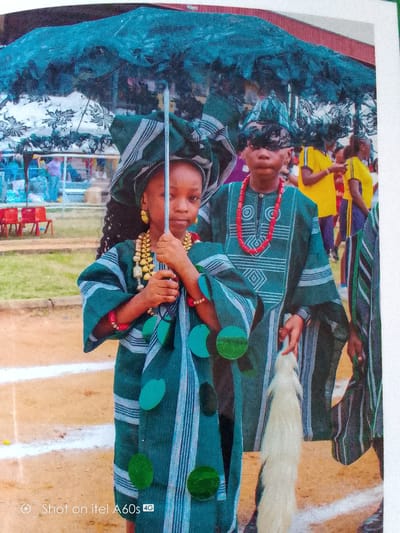 The image shows a group of children in Idimu, Nigeria, dressed in traditional attire. Two children are centrally located in the foreground. The girl in front is holding an umbrella with a silver shaft. She has dark hair, gold earrings, and beaded necklaces. The boy behind her is wearing a patterned green top and a red beaded necklace. Both are wearing matching headwraps. In the background, other people are visible, including individuals in yellow shirts. The setting appears to be outdoors with chairs in the background and what may be a running track on the ground. The bottom left corner of the image has the text "Shot on itel A60s 4G."