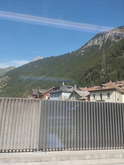 The image depicts a mountain landscape with a village in the foreground, situated in Göschenen, Switzerland. A ribbed, concrete barrier runs across the bottom of the frame, obscuring the lower portion of the scene and casting shadows. Behind the barrier, several houses with pitched roofs are visible, clustered together. The houses vary in color, with some featuring terracotta tiles and others lighter facades. A prominent church steeple is not discernible, but the architecture suggests a traditional alpine village. The mountain slopes behind the village are densely covered with coniferous trees, forming a dark green carpet that stretches towards the rocky, rugged peaks at the top of the image. The sky is a clear, bright blue, with a faint white streak running horizontally across it, possibly a reflection or a contrail. A tall, lattice-like telecommunications tower is positioned on the mountainside, adding a modern element to the otherwise natural and rustic setting. The overall impression is one of a serene, mountainous region undergoing development.