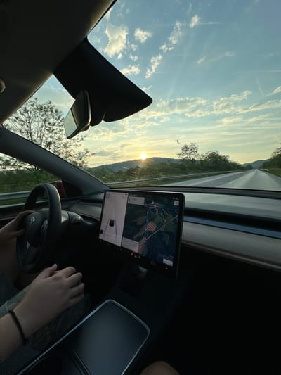 The image captures the interior of a Tesla vehicle looking out through the windshield at sunset in Dechkovtsi, Bulgaria. The sun, partially obscured by the horizon, is setting on the right side of the frame, casting an orange glow and lens flares across the windshield. The vehicle is on a paved two-lane road flanked by guardrails and lush green hills covered with trees and foliage. The sky above is a mix of clear blue and scattered white clouds, with faint contrails visible.

Inside the vehicle, a person's left hand is visible on the steering wheel, which features the Tesla logo. The central touchscreen display is active, showing a digital speedometer indicating 85 km/h, an estimated range of 67 km, and an ambient temperature of 18°C. The navigation system displays a map of a rural area with Cyrillic text, alongside a vehicle visualization indicating active driving assistance. The time displayed is 08:32. Below the screen, the cabin temperature is set to 21.5°C. Another person's right hand, wearing a black beaded bracelet, rests on the center console, which appears to have a textured, dark surface. A rearview mirror is visible mounted to the overhead console, partially reflecting the sky.
