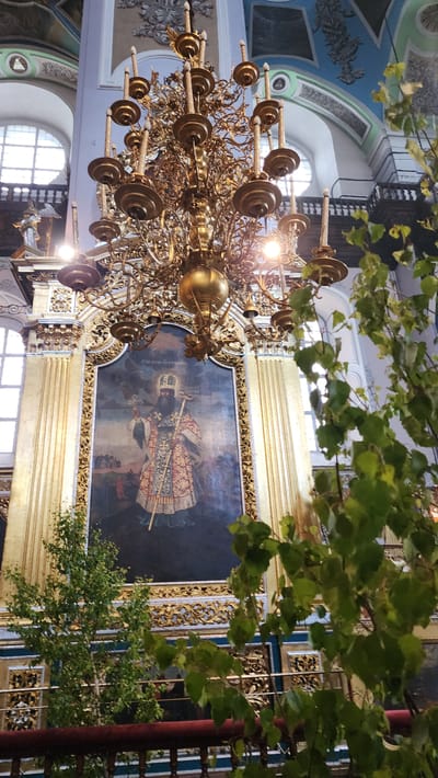 The image shows the interior of a church in Smolensk, Russia. A large, golden chandelier hangs in the foreground, with numerous arms and candle holders. Behind the chandelier, a large framed icon is mounted on the wall. The icon depicts a religious figure holding a staff and wearing elaborate vestments. The wall is painted with decorative designs in blue, gold, and white. Green foliage is present in the upper and lower right corners of the image. Windows can be seen in the background, providing natural light.