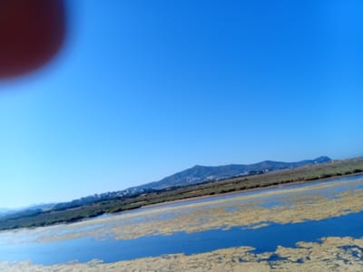 The image depicts a landscape in Martil, Morocco. In the foreground is a body of water covered with aquatic vegetation. In the midground, there is a strip of land with green vegetation and some scattered buildings. In the background, a mountain range is visible under a clear blue sky. A blurred red object is in the upper left corner.
