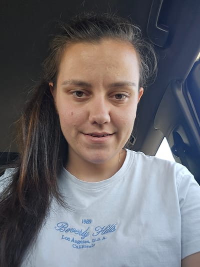 A woman with long, dark brown hair is pictured from the chest up, inside what appears to be a vehicle. She is wearing a light blue t-shirt with "1989 Beverly Hills Los Angeles, U.S.A. California" embroidered in blue script on the front. The woman is looking directly at the camera with a slight smile. Her face is slightly flushed, and her hair is pulled back into a ponytail on her left side. The background is dark, with a portion of the car's interior visible, including a sun visor and a window. The specific location in Staro Selo, Bulgaria, is not discernible from the image.