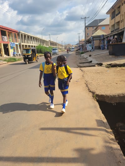 Two young girls, dressed in matching yellow and blue school uniforms with white socks and shoes, stand on a paved road. Their uniforms feature a crest on the left breast. The girl in the foreground has her left arm bent at the elbow, with her hand near her waist, and her right arm extended downwards. The girl behind her has both arms bent at the elbows, with her hands near her chest. Both girls have dark hair styled in braids.
The scene appears to be an outdoor street setting, possibly in Nigeria given the context provided. The road is made of asphalt and is lined with buildings on the left side of the frame. These buildings appear to be shops or residential structures, with awnings and signage visible. On the right side of the road, there is a raised earthen embankment. The sky is visible in the background, with scattered white clouds suggesting it is daytime and possibly sunny, as indicated by the clear shadows cast on the ground. A yellow and black motorized tricycle, commonly known as a "Keke Napep," is parked on the left side of the road, behind the girls. In the distance, there are other vehicles and more buildings. The overall impression is a typical street scene in a populated area. There is no discernible text in the image that can be read clearly.