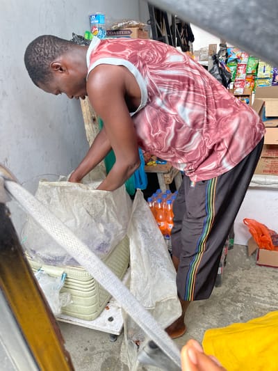 An African man, wearing a red and white marble-patterned tank top and black track pants with a multicolored stripe down the side, is rummaging through a large, opaque white plastic bag. He is bent over, his arms inside the bag, and his face is obscured as he looks down. The man is standing on a concrete floor in what appears to be an outdoor market or shop setting in Ilado, Nigeria. Behind him, shelves are stocked with various packaged goods, including drinks in plastic bottles, bags of snacks, and cardboard boxes. A light-colored ribbed plastic basket is visible beneath the white bag the man is accessing. To the left, a thick, white, ribbed hose or tube is prominently featured in the foreground, obscuring part of the man and the environment. A yellow fabric is visible in the lower right corner of the image. The background wall is a rough, light gray surface.