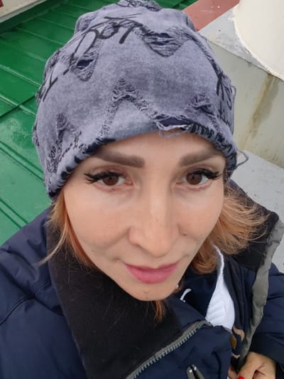 A woman with reddish-blonde hair, wearing a gray knit hat with ripped detailing and a dark blue jacket over a white top, is taking a selfie. She has on false eyelashes and pink lipstick. The background consists of a green metal surface on the left and a light-colored cylindrical object on the right, with a gray, possibly concrete surface in between. The image appears to be taken outdoors. There is no information available in the image to determine the location is Ratibor, Russia.