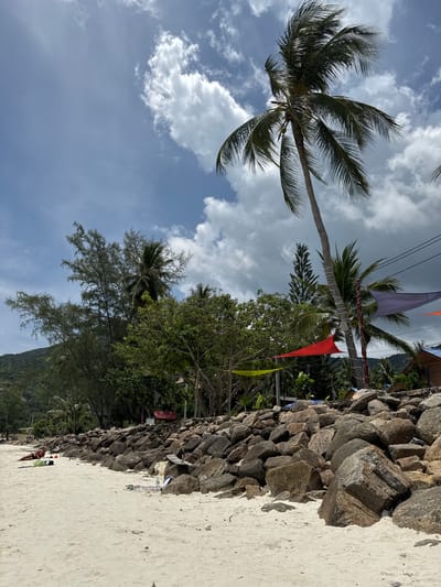 The image shows a beach scene in Baan Chaloklum, Thailand. A palm tree is on the left side of the image, with white sand in the foreground and a rock wall running along the middle right side of the scene. Lush trees and vegetation are positioned between the palm tree and rock wall and, further in the background, a glimpse of a mountain can be seen. Several colorful flags are strung between trees. The sky is bright with scattered clouds.
