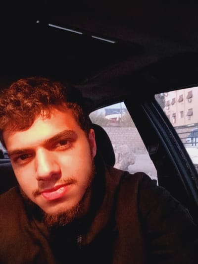 The image depicts a young adult male situated within the dark interior of a vehicle, appearing to take a self-portrait. He has dark, curly hair, a beard, and wears a dark, collared jacket with a zipper. His gaze is directed forward, and his tongue is slightly visible between his lips. A strong light source from the front-right illuminates the right side of his face and upper body, while the left side remains in shadow. Through the vehicle's windows, an urban exterior of Kafr Sousa Municipality, Syria, is visible. On the right, a multi-story, light-colored building with multiple windows and several protruding exterior units, possibly air conditioning units, is present. Further in the background, additional structures and a light-colored wall or rocky formation can be discerned. The vehicle's dark overhead console is partially visible at the top of the frame.
