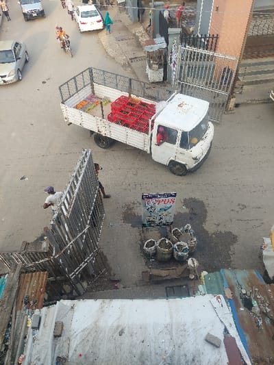 A truck filled with red baskets sits on a street, alongside a small white van. Adjacent to them are multiple woven baskets, possibly containing goods for sale. A sign stands nearby. The street scene includes several cars and pedestrians. Overhanging structures and buildings border the road, creating an urban setting. The image appears to be taken from above, possibly from a higher building.